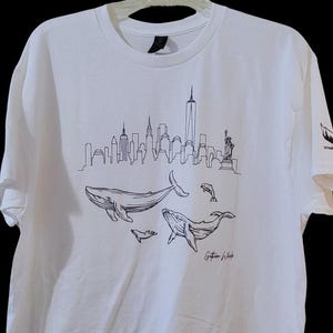 Puede incluir: Camiseta blanca con un dibujo lineal negro del horizonte de Nueva York, ballenas y delfines. La camiseta incluye el texto "Gotham Whale" en una fuente de escritura. La camiseta est&aacute; colgada en una percha blanca.