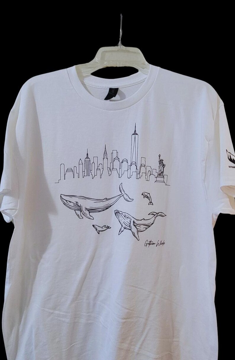 Puede incluir: Camiseta blanca con un dibujo lineal negro del horizonte de Nueva York, ballenas y delfines. La camiseta incluye el texto "Gotham Whale" en una fuente de escritura. La camiseta est&aacute; colgada en una percha blanca.