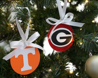 May include: Christmas tree ornaments featuring college team logos. One ornament is orange with a white "T", the other is red with a black and white "G". Both are round and have white ribbon for hanging.