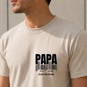 Puede incluir: Camiseta beige con la palabra "PAPA" en letras negras en negrita. Debajo, un diseño de código de barras con el texto "ETS. 22-26" y los nombres "Anna ❤ Noah".