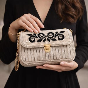 May include: A beige crossbody bag with a gold chain strap and clasp. The bag features a black floral design on the flap and a textured, ribbed pattern on the body. The bag is held by a person wearing a black dress.