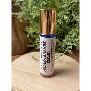 Peut inclure: Un flacon d'huile parfumée à bille avec un bouchon doré et une étiquette blanche. L'étiquette indique "Scent is inspired by Ariana Grande Cloud Luxury Fragrance Oil". Le flacon est posé sur un fond de verdure et une surface en bois.