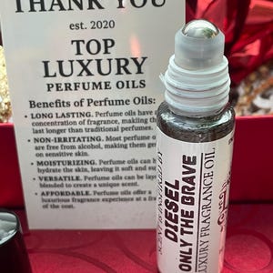 May include: A perfume oil bottle with a silver rollerball top and a white label. The label reads "DIESEL ONLY THE BRAVE LUXURY FRAGRANCE OIL". A card behind the bottle says "THANK YOU" and "TOP LUXURY PERFUME OILS".
