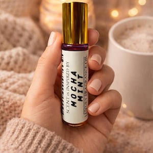Op de afbeelding: Een kleine, cilindrische glazen fles luxe geurolie met een gouden dop wordt in een hand gehouden. Het etiket luidt "Scent is inspired by Mocha Mint". De achtergrond omvat een brandende kaars en een mok.
