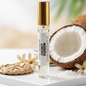 Puede incluir: Un frasco roll-on de vidrio transparente con tapa dorada, etiquetado como "Scent Inspired by Coconut Passion Luxury Fragrance Oil". El frasco est&aacute; sobre una superficie blanca con media coco, flores y cristales de sal.