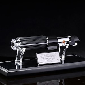 Könnte beinhalten: Eine Darth Vader Lichtschwert-Replik aus Star Wars, präsentiert auf einer schwarzen Basis mit einem transparenten Acrylständer. Das Lichtschwert hat ein silbernes und schwarzes Design mit detaillierten Merkmalen. Eine kleine Plakette trägt die Aufschrift "Darth Vader Lightsaber".