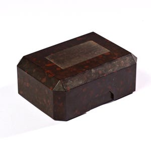 Könnte beinhalten: Eine rechteckige Box im antiken Stil mit dunkelbrauner, gesprenkelter Oberfläche. Die Box hat einen Klappdeckel mit einem rechteckigen, metallischen Akzent. Die Kanten der Box sind facettiert und verleihen dem Design ein einzigartiges geometrisches Detail.