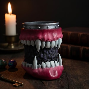 May include: A novelty mug shaped like a monstrous mouth, featuring large white fangs and a dark purple interior. The rim is silver, and the lips are painted a deep red. A lit candle and antique key are in the background.