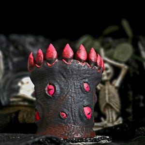 Może przedstawiać: Czarny i czerwony pojemnik dekoracyjny o tematyce Halloween. Pojemnik ma teksturowaną powierzchnię z czerwonymi akcentami i koroną. Pojemnik ma kilka czerwonych detali w kształcie oka. Pojemnik stoi na czarnej powierzchni.