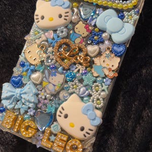 Peut inclure: Une coque de t&eacute;l&eacute;phone transparente orn&eacute;e de personnages Hello Kitty, de n&oelig;uds et de divers ornements bleus et blancs. La coque pr&eacute;sente l'inscription "BIG LEO" en lettres dor&eacute;es et un c&oelig;ur dor&eacute; avec le mot "LOVE".