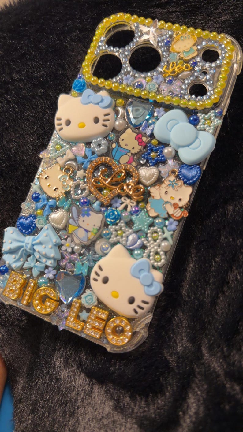Peut inclure: Une coque de t&eacute;l&eacute;phone transparente orn&eacute;e de personnages Hello Kitty, de n&oelig;uds et de divers ornements bleus et blancs. La coque pr&eacute;sente l'inscription "BIG LEO" en lettres dor&eacute;es et un c&oelig;ur dor&eacute; avec le mot "LOVE".