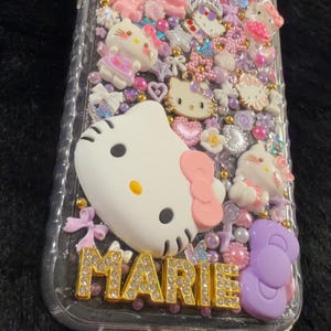 Peut inclure: Coque de t&eacute;l&eacute;phone transparente orn&eacute;e de personnages Hello Kitty, de n&oelig;uds et de divers ornements roses, blancs et violets. Le nom "MARIE" est &eacute;crit en lettres dor&eacute;es en bas.
