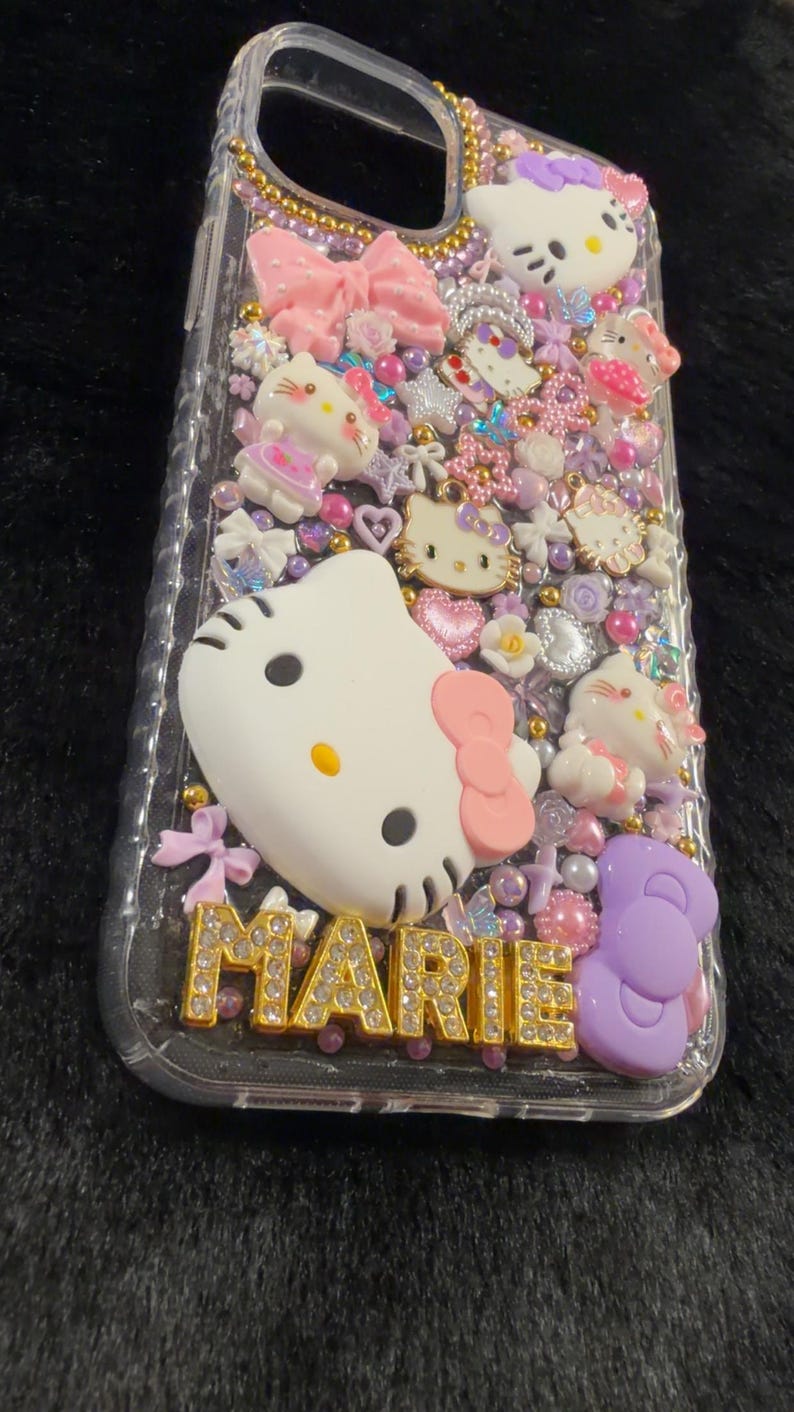 Peut inclure: Coque de t&eacute;l&eacute;phone transparente orn&eacute;e de personnages Hello Kitty, de n&oelig;uds et de divers ornements roses, blancs et violets. Le nom "MARIE" est &eacute;crit en lettres dor&eacute;es en bas.