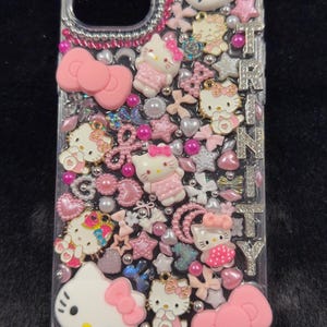 Peut inclure: Coque de t&eacute;l&eacute;phone transparente orn&eacute;e de personnages Hello Kitty roses et blancs, de n&oelig;uds, d'&eacute;toiles et de perles. Le nom "TRINITY" est &eacute;pel&eacute; en lettres argent&eacute;es. L'&eacute;tui est orn&eacute; de strass roses.