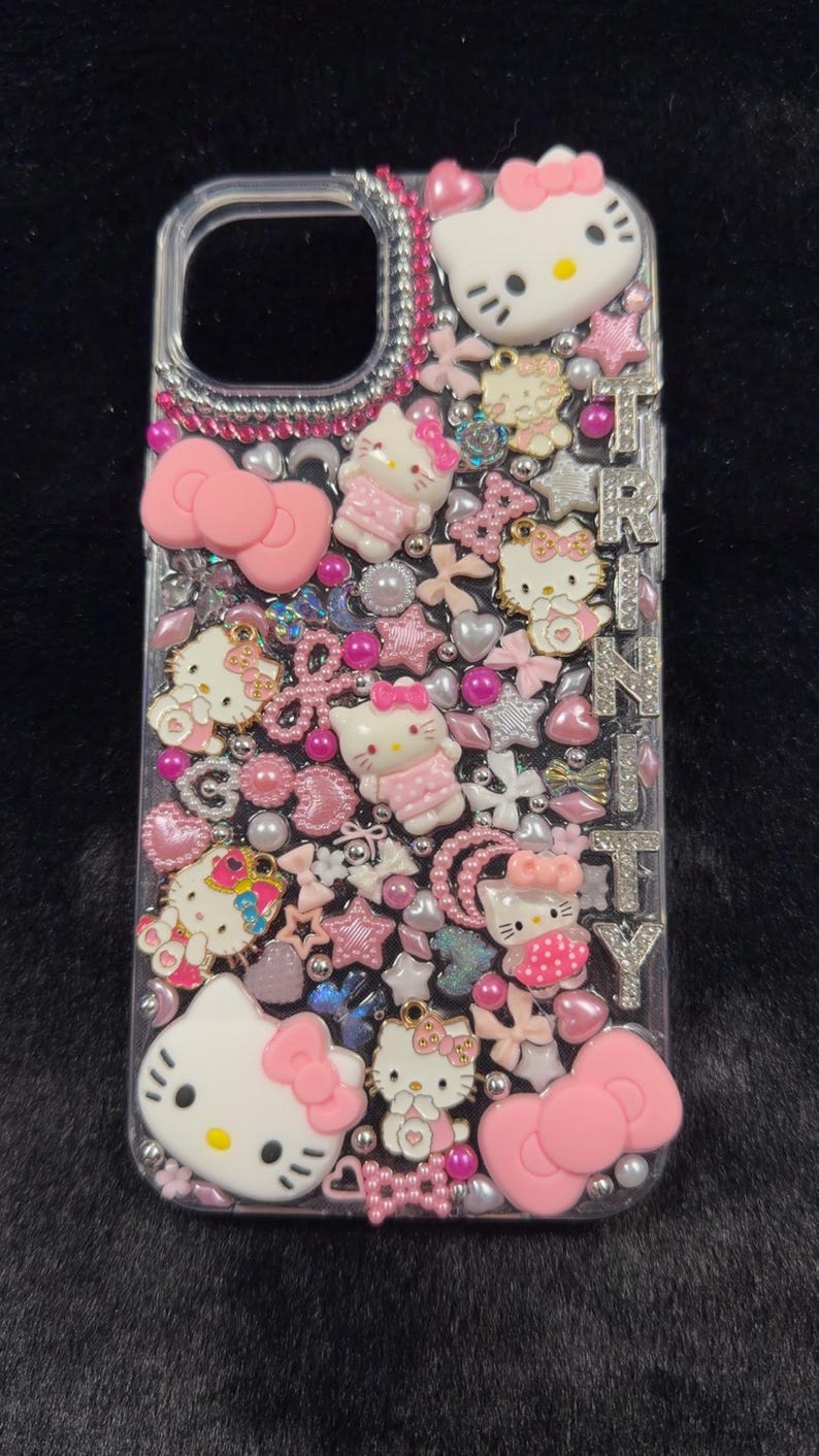 Peut inclure: Coque de t&eacute;l&eacute;phone transparente orn&eacute;e de personnages Hello Kitty roses et blancs, de n&oelig;uds, d'&eacute;toiles et de perles. Le nom "TRINITY" est &eacute;pel&eacute; en lettres argent&eacute;es. L'&eacute;tui est orn&eacute; de strass roses.
