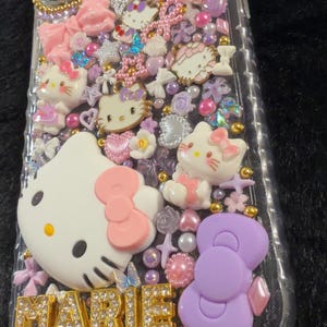 Peut inclure: Coque de t&eacute;l&eacute;phone transparente d&eacute;cor&eacute;e de figurines Hello Kitty, de n&oelig;uds et d'autres ornements roses, blancs et violets. Le nom "MARIE" est &eacute;pel&eacute; en lettres dor&eacute;es. L'&eacute;tui est recouvert de petits &eacute;l&eacute;ments d&eacute;coratifs.