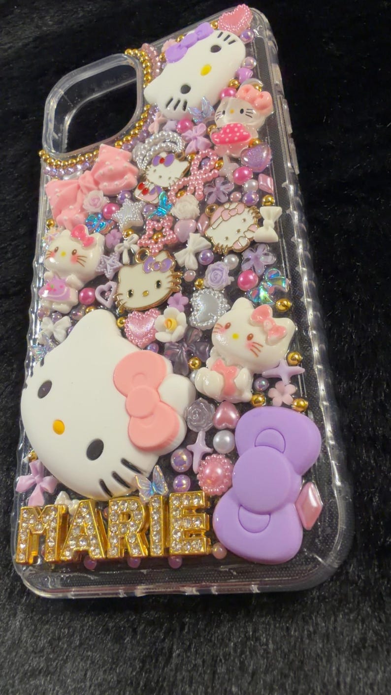 Peut inclure: Coque de t&eacute;l&eacute;phone transparente d&eacute;cor&eacute;e de figurines Hello Kitty, de n&oelig;uds et d'autres ornements roses, blancs et violets. Le nom "MARIE" est &eacute;pel&eacute; en lettres dor&eacute;es. L'&eacute;tui est recouvert de petits &eacute;l&eacute;ments d&eacute;coratifs.