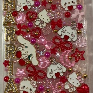 Peut inclure: Une coque de t&eacute;l&eacute;phone transparente orn&eacute;e d'&eacute;l&eacute;ments roses, rouges et dor&eacute;s. Elle pr&eacute;sente des personnages Hello Kitty et Cinnamoroll, des c&oelig;urs, des n&oelig;uds et le nom "NARAH" en lettres dor&eacute;es. Le design est compos&eacute; de perles et de breloques.