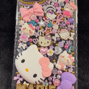Peut inclure: Coque de t&eacute;l&eacute;phone transparente orn&eacute;e de figurines Hello Kitty, de n&oelig;uds, de c&oelig;urs et de perles roses, violettes et blanches. Le nom "MARIE" est &eacute;pel&eacute; en lettres dor&eacute;es en bas.