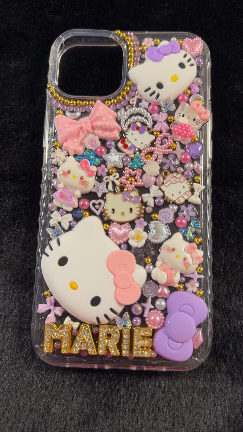 Peut inclure: Coque de t&eacute;l&eacute;phone transparente orn&eacute;e de figurines Hello Kitty, de n&oelig;uds, de c&oelig;urs et de perles roses, violettes et blanches. Le nom "MARIE" est &eacute;pel&eacute; en lettres dor&eacute;es en bas.