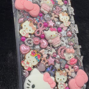 Peut inclure: Coque de t&eacute;l&eacute;phone transparente orn&eacute;e de n&oelig;uds roses, de figurines Hello Kitty et de divers embellissements roses et argent&eacute;s. Le mot "TRINITY" est &eacute;pel&eacute; en lettres argent&eacute;es sur le c&ocirc;t&eacute;. La coque est recouverte de strass et de perles.