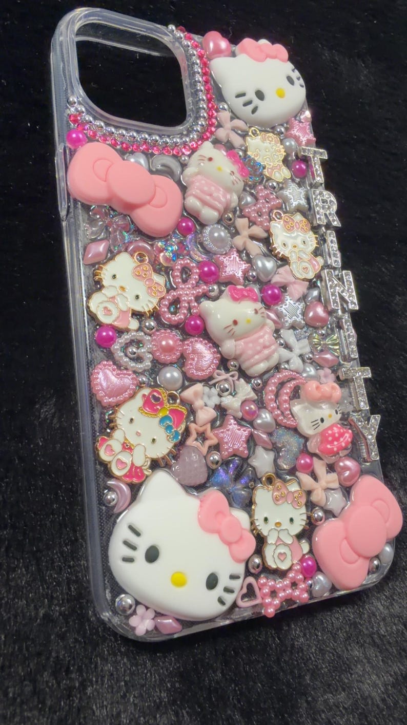 Peut inclure: Coque de t&eacute;l&eacute;phone transparente orn&eacute;e de n&oelig;uds roses, de figurines Hello Kitty et de divers embellissements roses et argent&eacute;s. Le mot "TRINITY" est &eacute;pel&eacute; en lettres argent&eacute;es sur le c&ocirc;t&eacute;. La coque est recouverte de strass et de perles.