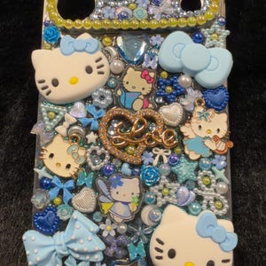 Peut inclure: Coque de t&eacute;l&eacute;phone transparente orn&eacute;e de personnages Hello Kitty, de n&oelig;uds et d'&eacute;l&eacute;ments d&eacute;coratifs bleus, blancs et dor&eacute;s. La coque porte l'inscription "BIG LEO" en lettres dor&eacute;es et strass.