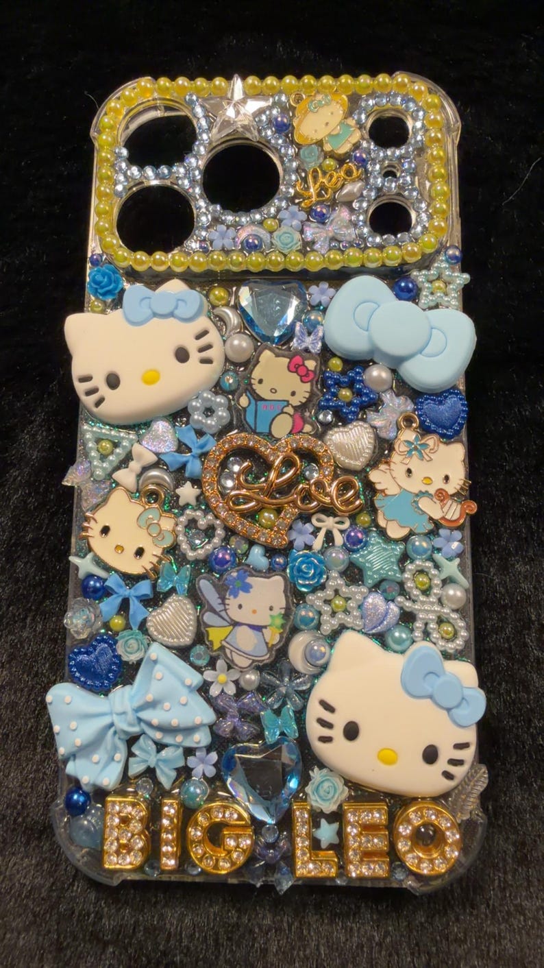 Peut inclure: Coque de t&eacute;l&eacute;phone transparente orn&eacute;e de personnages Hello Kitty, de n&oelig;uds et d'&eacute;l&eacute;ments d&eacute;coratifs bleus, blancs et dor&eacute;s. La coque porte l'inscription "BIG LEO" en lettres dor&eacute;es et strass.