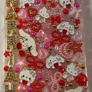 Peut inclure: Une coque de t&eacute;l&eacute;phone rose orn&eacute;e de Hello Kitty, Cinnamoroll, de c&oelig;urs, de n&oelig;uds et d'accents dor&eacute;s. Le nom "MARIAH" est &eacute;pel&eacute; en lettres dor&eacute;es. L'&eacute;tui est orn&eacute; de divers embellissements roses et rouges.
