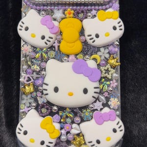 Peut inclure: Coque de t&eacute;l&eacute;phone transparente orn&eacute;e de visages Hello Kitty, de n&oelig;uds violets et de divers ornements. L'&eacute;tui pr&eacute;sente une couronne jaune et est orn&eacute; de strass, de perles et d'&eacute;l&eacute;ments floraux. Les visages Hello Kitty ont des nez jaunes et des moustaches noires.