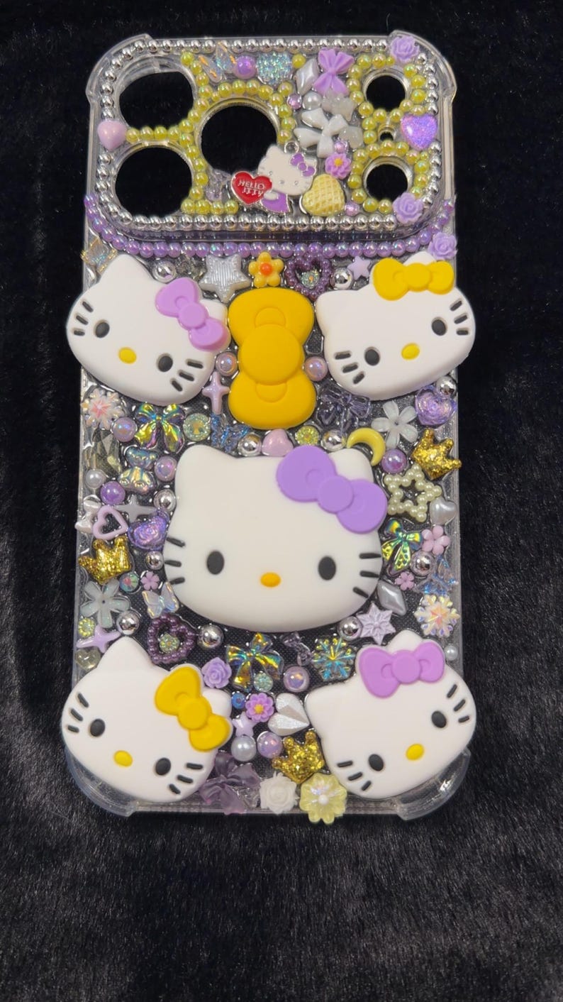 Peut inclure: Coque de t&eacute;l&eacute;phone transparente orn&eacute;e de visages Hello Kitty, de n&oelig;uds violets et de divers ornements. L'&eacute;tui pr&eacute;sente une couronne jaune et est orn&eacute; de strass, de perles et d'&eacute;l&eacute;ments floraux. Les visages Hello Kitty ont des nez jaunes et des moustaches noires.