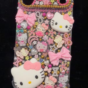 Peut inclure: Une coque de t&eacute;l&eacute;phone transparente orn&eacute;e de n&oelig;uds roses, de figurines Hello Kitty, de fleurs et de perles dor&eacute;es. L'&eacute;tui a une bordure dor&eacute;e et est orn&eacute; d'accents roses et blancs. Le design a un th&egrave;me mignon et girly.