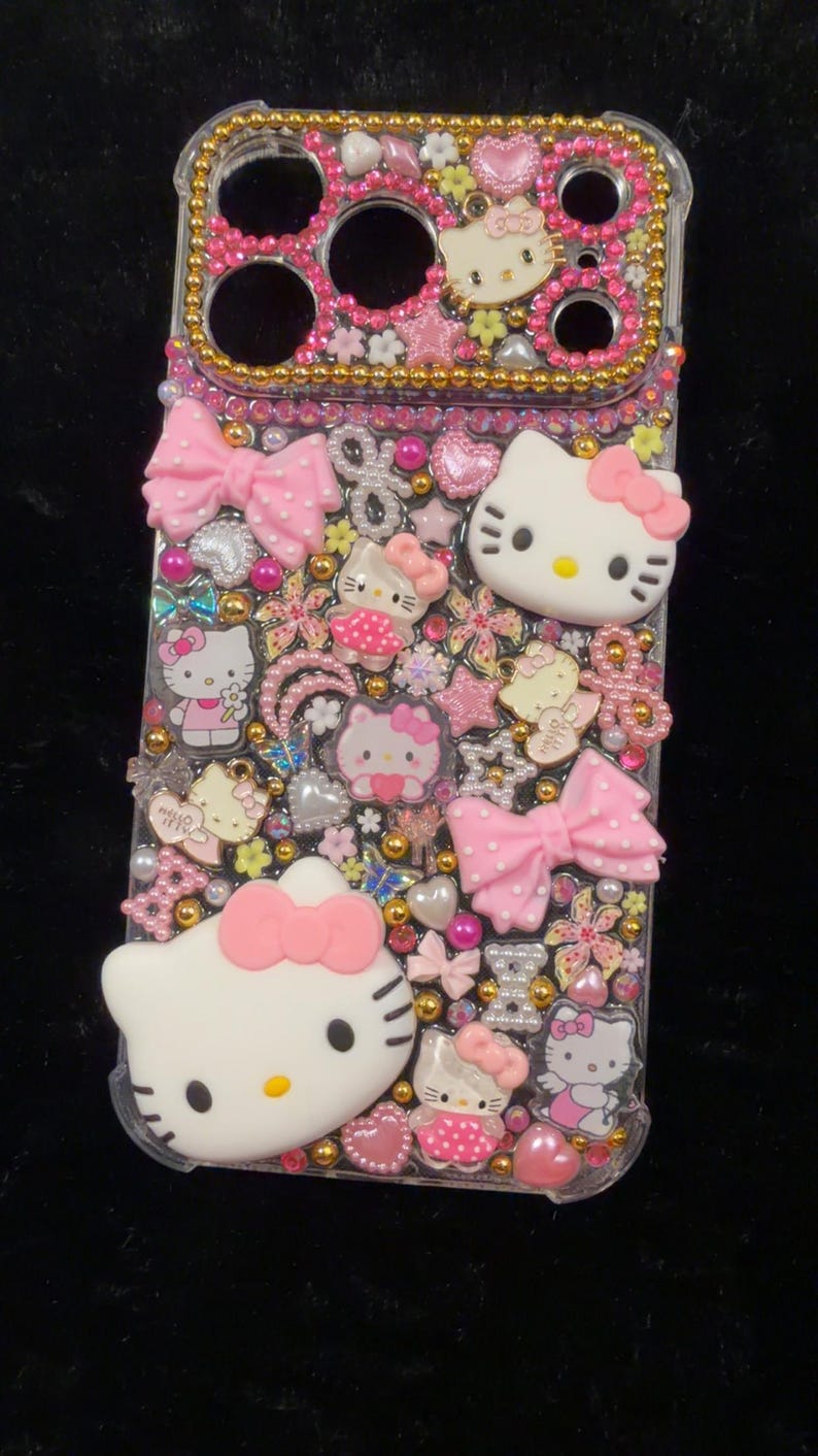 Peut inclure: Une coque de t&eacute;l&eacute;phone transparente orn&eacute;e de n&oelig;uds roses, de figurines Hello Kitty, de fleurs et de perles dor&eacute;es. L'&eacute;tui a une bordure dor&eacute;e et est orn&eacute; d'accents roses et blancs. Le design a un th&egrave;me mignon et girly.