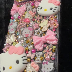 Peut inclure: Coque de t&eacute;l&eacute;phone transparente orn&eacute;e de n&oelig;uds roses, de figurines Hello Kitty et de divers ornements. L'&eacute;tui pr&eacute;sente un bord dor&eacute; et est orn&eacute; d'accents roses, dor&eacute;s et nacr&eacute;s, cr&eacute;ant un design ludique et d&eacute;coratif.