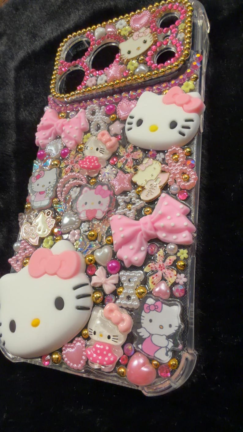 Peut inclure: Coque de t&eacute;l&eacute;phone transparente orn&eacute;e de n&oelig;uds roses, de figurines Hello Kitty et de divers ornements. L'&eacute;tui pr&eacute;sente un bord dor&eacute; et est orn&eacute; d'accents roses, dor&eacute;s et nacr&eacute;s, cr&eacute;ant un design ludique et d&eacute;coratif.