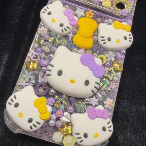 Peut inclure: Coque de t&eacute;l&eacute;phone transparente orn&eacute;e de figurines Hello Kitty blanches avec des n&oelig;uds violets et jaunes. L'&eacute;tui est orn&eacute; de perles, de fleurs et d'autres &eacute;l&eacute;ments d&eacute;coratifs dans des tons violets, jaunes et dor&eacute;s.