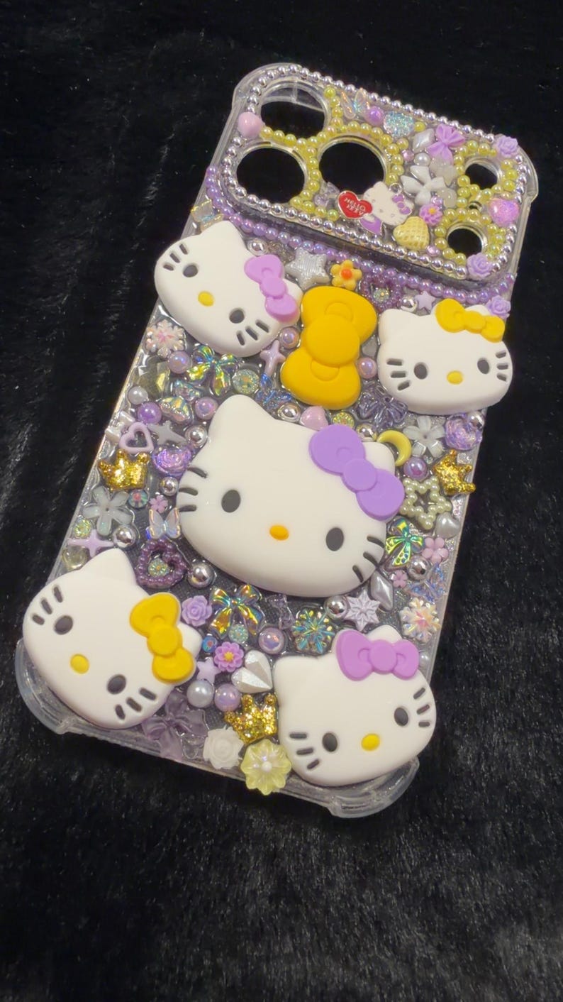 Peut inclure: Coque de t&eacute;l&eacute;phone transparente orn&eacute;e de figurines Hello Kitty blanches avec des n&oelig;uds violets et jaunes. L'&eacute;tui est orn&eacute; de perles, de fleurs et d'autres &eacute;l&eacute;ments d&eacute;coratifs dans des tons violets, jaunes et dor&eacute;s.