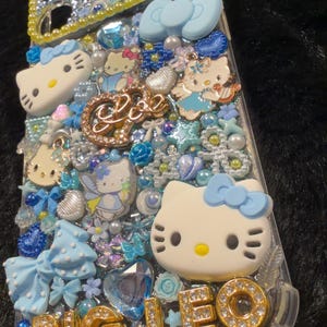 Peut inclure: Coque de t&eacute;l&eacute;phone transparente d&eacute;cor&eacute;e de figurines Hello Kitty, de n&oelig;uds bleus et d'ornements scintillants. La coque pr&eacute;sente le texte "BIG LEO" en lettres dor&eacute;es et est orn&eacute;e de divers bijoux bleus et transparents.