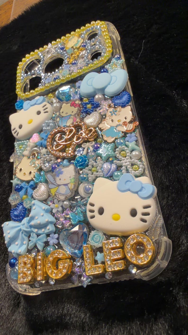 Peut inclure: Coque de t&eacute;l&eacute;phone transparente d&eacute;cor&eacute;e de figurines Hello Kitty, de n&oelig;uds bleus et d'ornements scintillants. La coque pr&eacute;sente le texte "BIG LEO" en lettres dor&eacute;es et est orn&eacute;e de divers bijoux bleus et transparents.