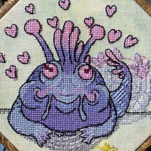 May include: Cross-stitch artwork of a purple creature with pink details and hearts, framed in a wooden hoop. The background is light green. The text "2024 Sternini / Ellen Bottini" is at the bottom. The frame has a decorative border. A unique piece of handmade art.