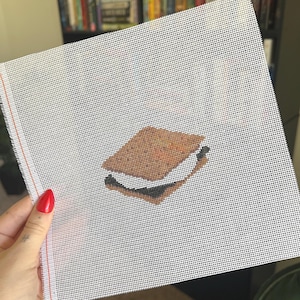 May include: A cross-stitch canvas featuring a s'more design. The s'more is depicted with a brown graham cracker, a melted marshmallow, and a dark chocolate square. The canvas is white with a grid pattern, and the edges are unfinished.