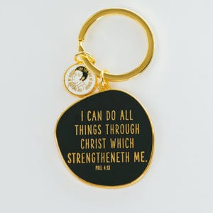 May include: Gold-tone keychain with a black pendant featuring the text "I CAN DO ALL THINGS THROUGH CHRIST WHICH STRENGTHENETH ME." A smaller round charm with a portrait is also attached. The keychain is a religious accessory.