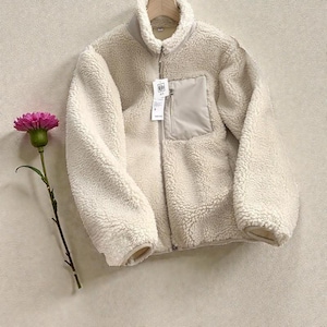 May include: A cream-coloured fleece jacket with a zip-up front and a small pocket on the chest. The jacket has a high collar and is hanging on a wooden hanger. A pink flower with a green stem is on the left side of the jacket.