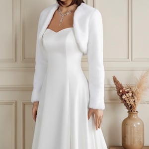 May include: A white faux fur bolero jacket with long sleeves, worn over a white wedding dress. The jacket has a soft, fluffy texture. The dress has a sweetheart neckline and a flowing skirt. A silver necklace is visible.