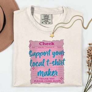 Könnte beinhalten: Cremefarbenes T-Shirt mit einem rosa und blauen Aufdruck "Support your local t-shirt maker". Eine goldene Stern-Anhänger-Halskette und ein brauner Hut sind ebenfalls im Bild.