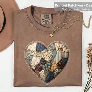 Peut inclure: Un t-shirt marron avec un motif de cœur patchwork imprimé. Le cœur est composé de divers motifs de tissu dans des tons de bleu, marron et crème. Un collier doré avec un pendentif étoile est posé sur le t-shirt. Le texte "Printed Patchwork Design" est visible.