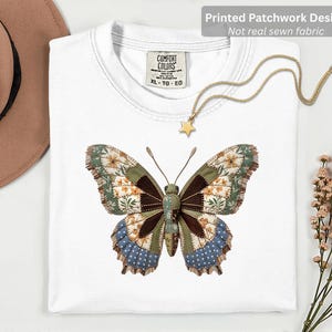 Könnte beinhalten: Weißes T-Shirt mit einem großen Schmetterlingsdesign. Der Schmetterling hat ein Patchwork-Design mit floralen, braunen und blauen Mustern. Eine goldene Halskette mit Sternanhänger liegt auf dem Shirt. Das Shirt hat ein Comfort Colors-Etikett.