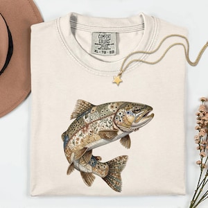 May include: Cream-coloured Comfort Colors XL t-shirt featuring a detailed illustration of a trout with floral and botanical patterns. A gold star pendant necklace and a brown hat are also visible. The shirt has a round neckline.