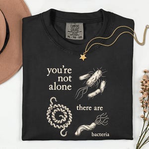 以下が含まれることがあります： 「you're not alone there are bacteria」の文字とバクテリアのイラストが描かれた黒いTシャツ。金の星のペンダントネックレスとベージュの帽子も画像に写っています。