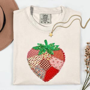 Puede incluir: Camiseta Comfort Colors color crema con un diseño de fresa de patchwork. La fresa está hecha de varias telas estampadas en rojo, rosa y blanco. Un collar con colgante de estrella dorada y un sombrero marrón también están en la imagen. La etiqueta dice XL-TG-EG.